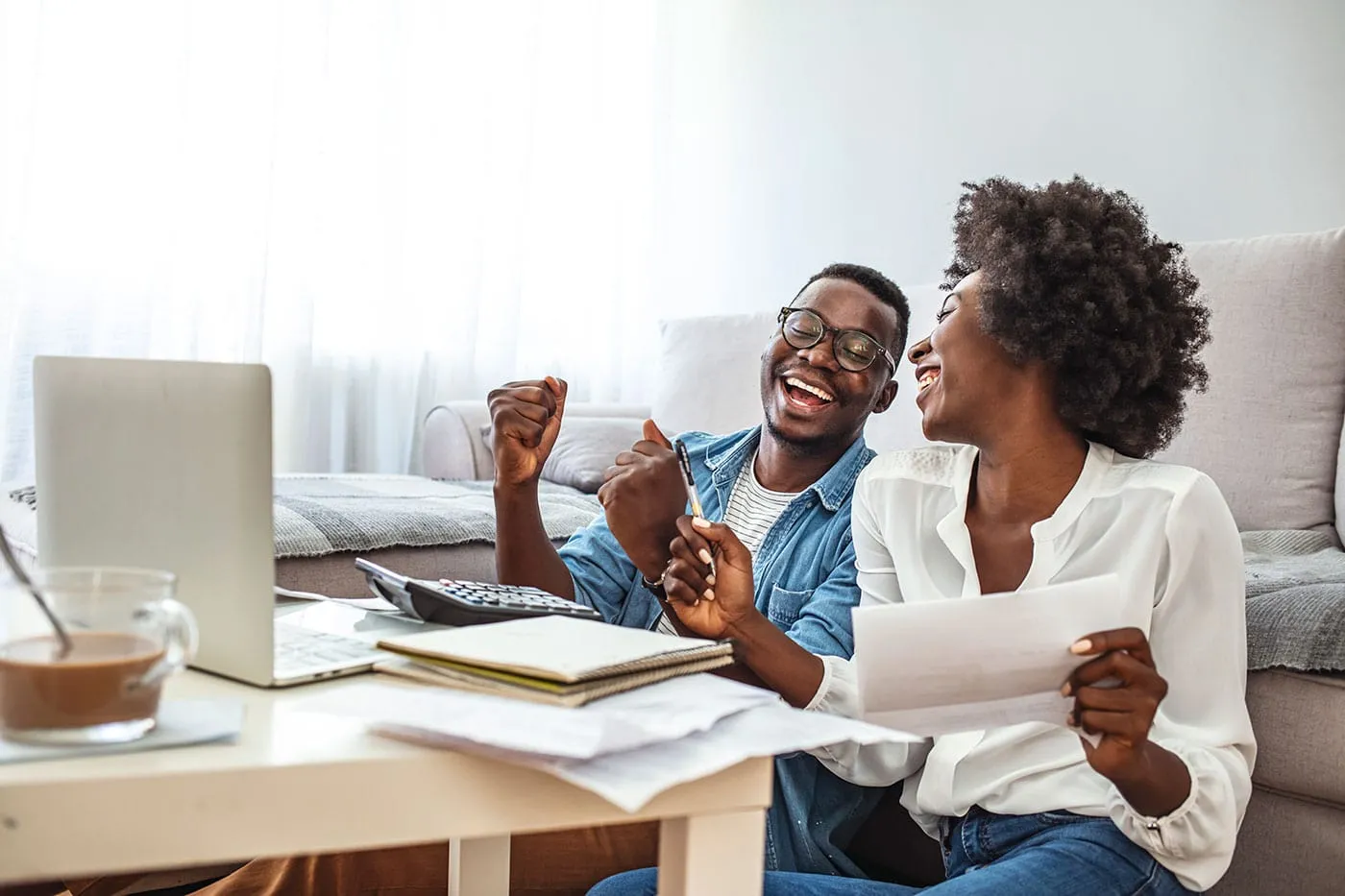 A couple sit on the living room floor while smiling at each other with their laptop and calculator on the table.