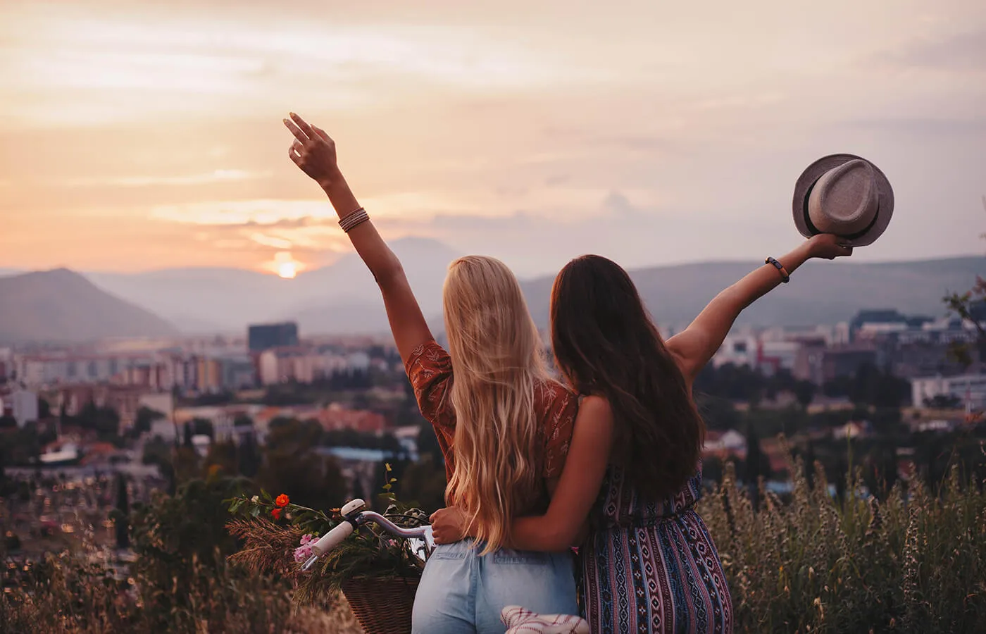 Two women standing on a cliff, facing the sunset and embracing.