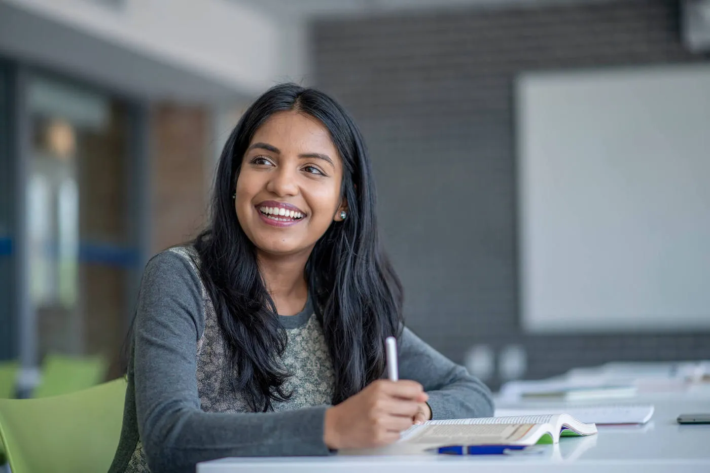 A college student wearing a gray shirt smiles while taking notes on her textbook.