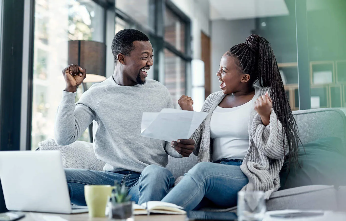 A couple sitting on the couch smiles and cheers together while holding documents.