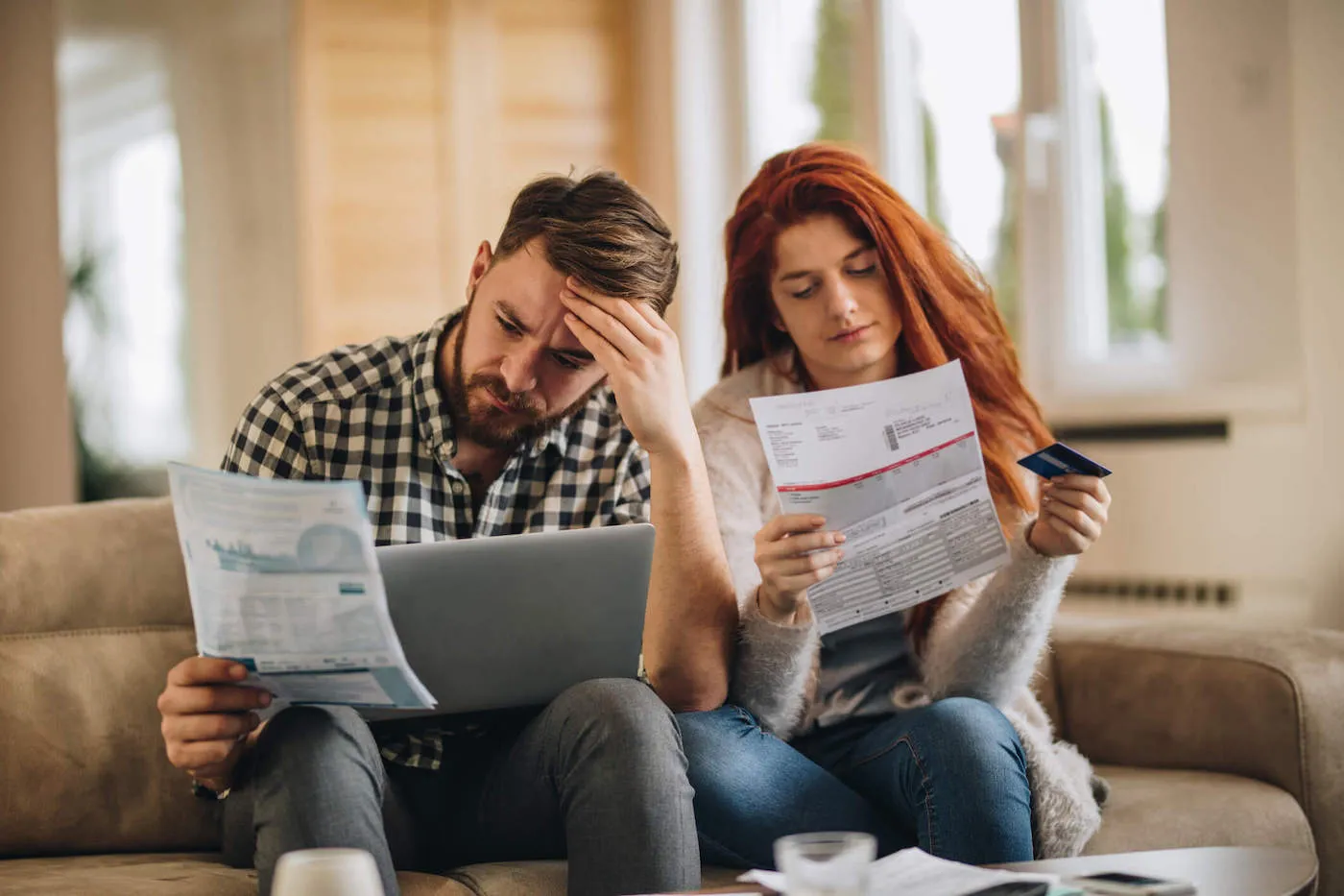 A couple frown while looking at bills as they sit on the couch.