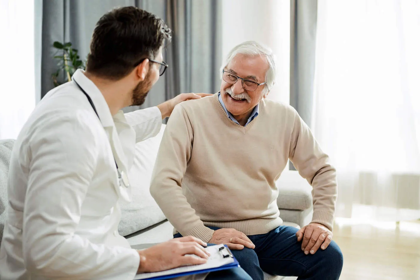 A doctor and an elderly patient smile at each other while sitting on the couch.