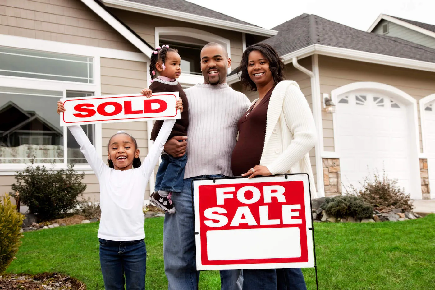 A family of four are smiling on the lawn of their new home with one of their daughter holding up a "Sold" sign.