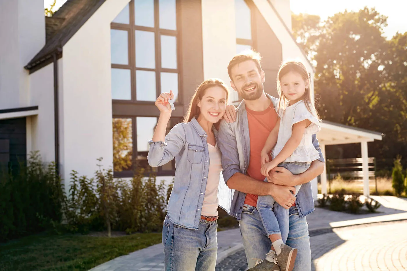 A family of three smiles outside their new home while the mother holds the house keys and the father holds up his daughter.