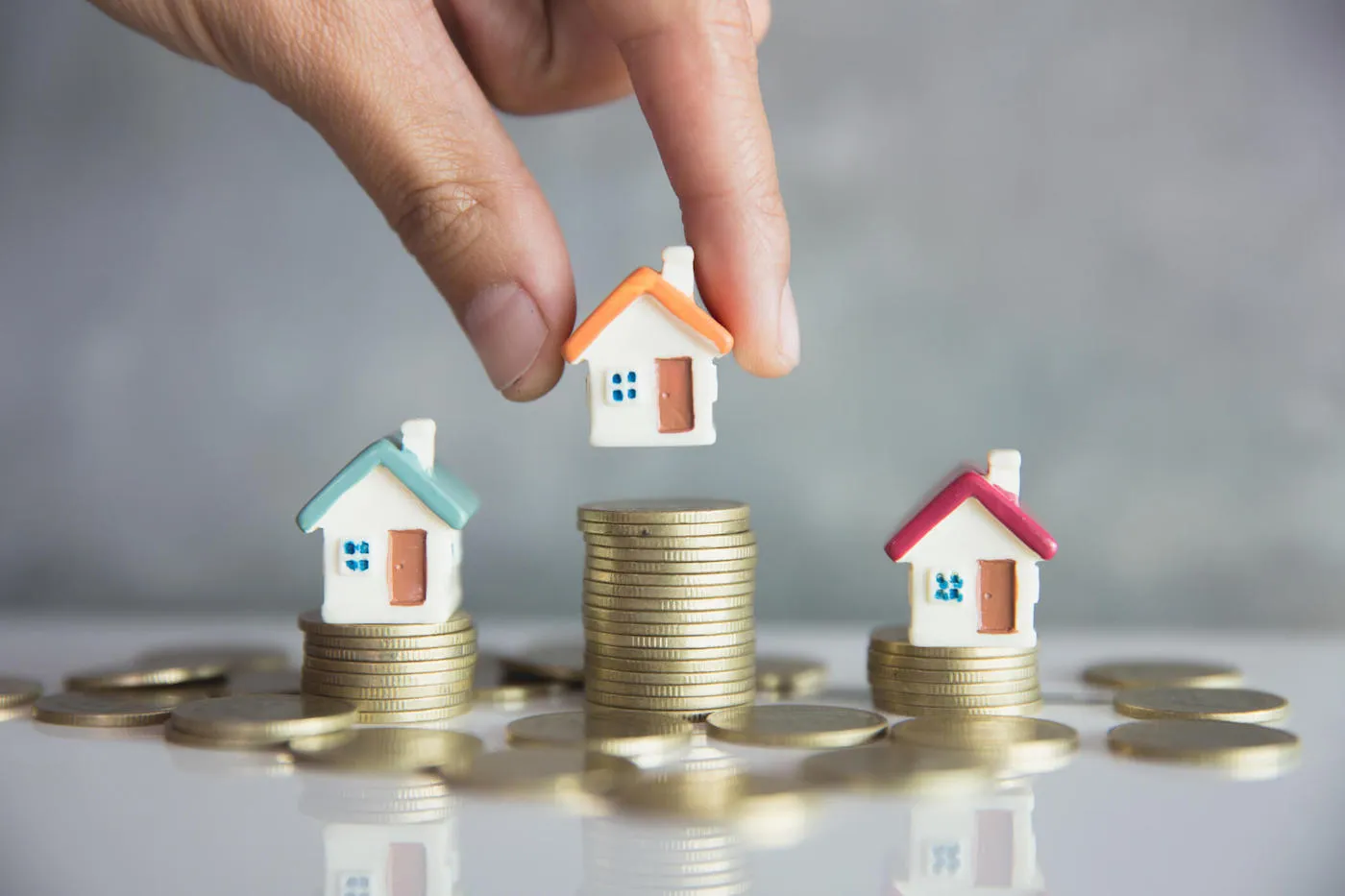 A hand holds one of three miniature house models which are placed on top of a stack of coins.