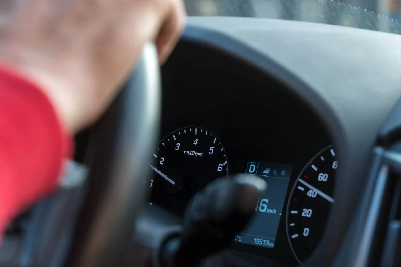 A hand hold the steering wheel of a car next to the car's dashboard.