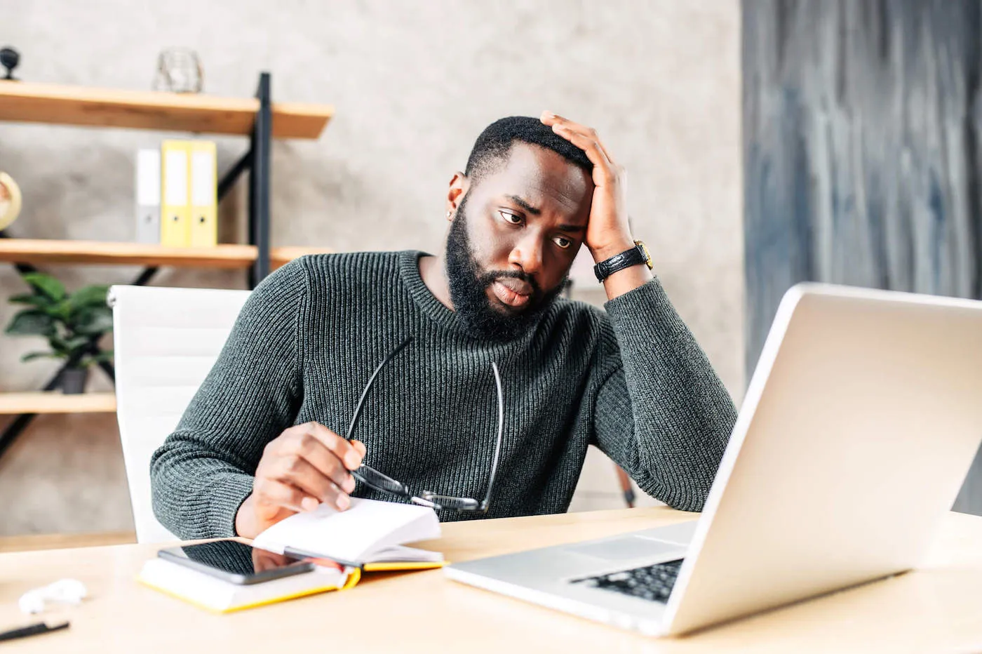 A man wearing a gray sweater has his hand to his head while he sits down looking at his laptop computer.