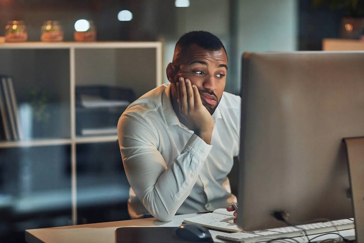 A man wearing white shirt is leaning on his hand as he looks at his computer screen.