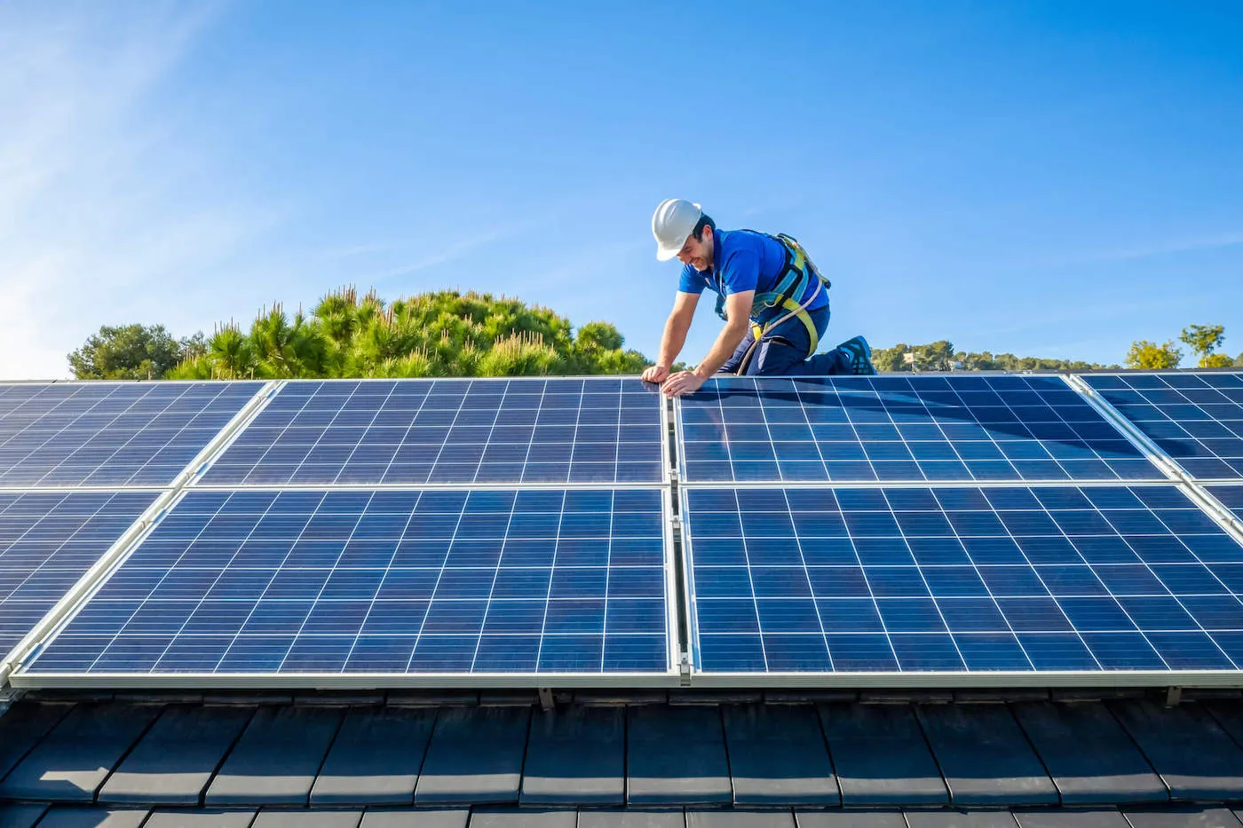 A man wearing a white hard hat installs solar panels on a roof of a home.