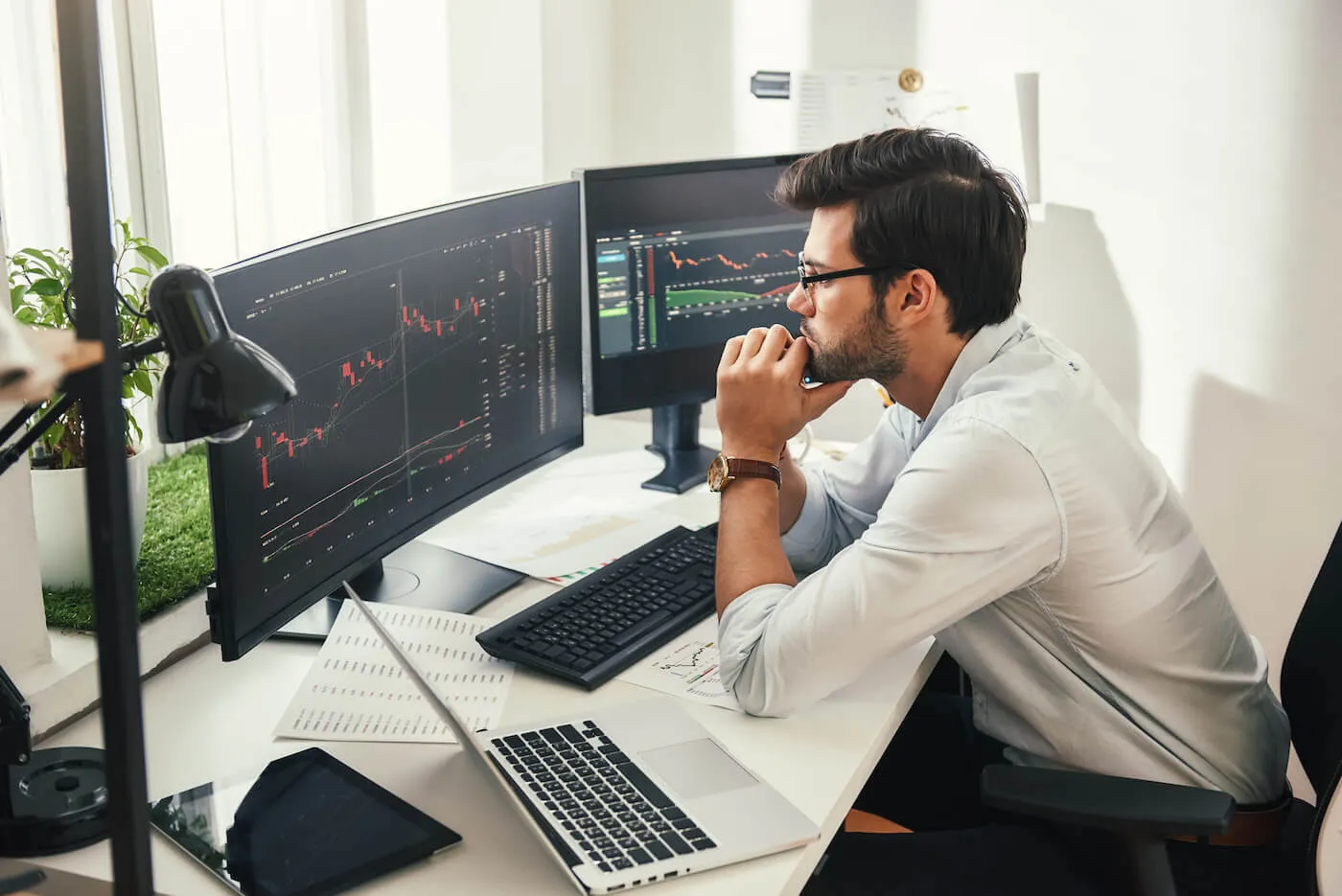 A man is at his work desk looking at two computer monitors that are showing stock graphs.