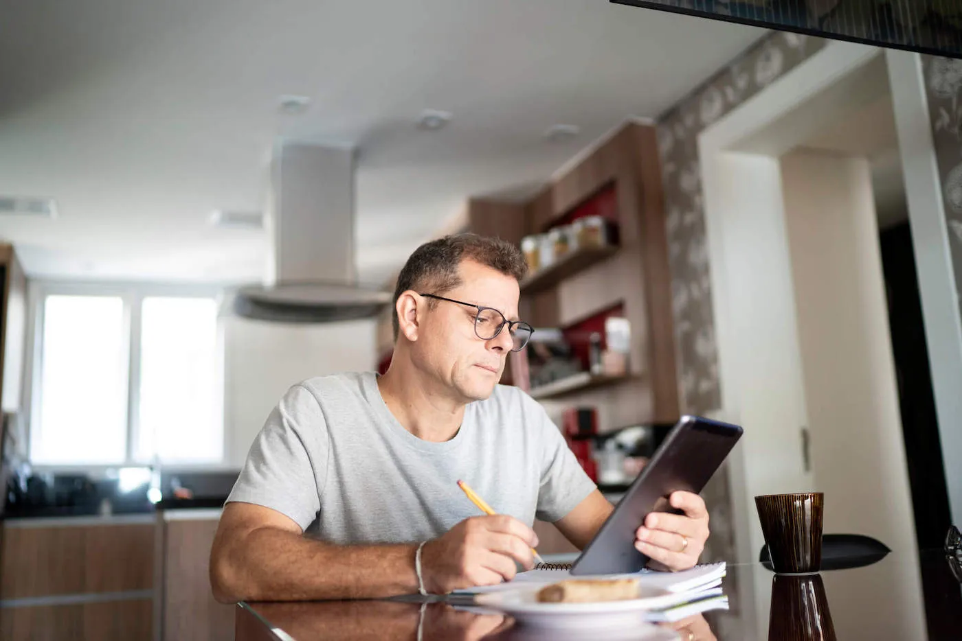 A man wearing a gray shirt and glasses looks at his tablet while writing on his notebook.