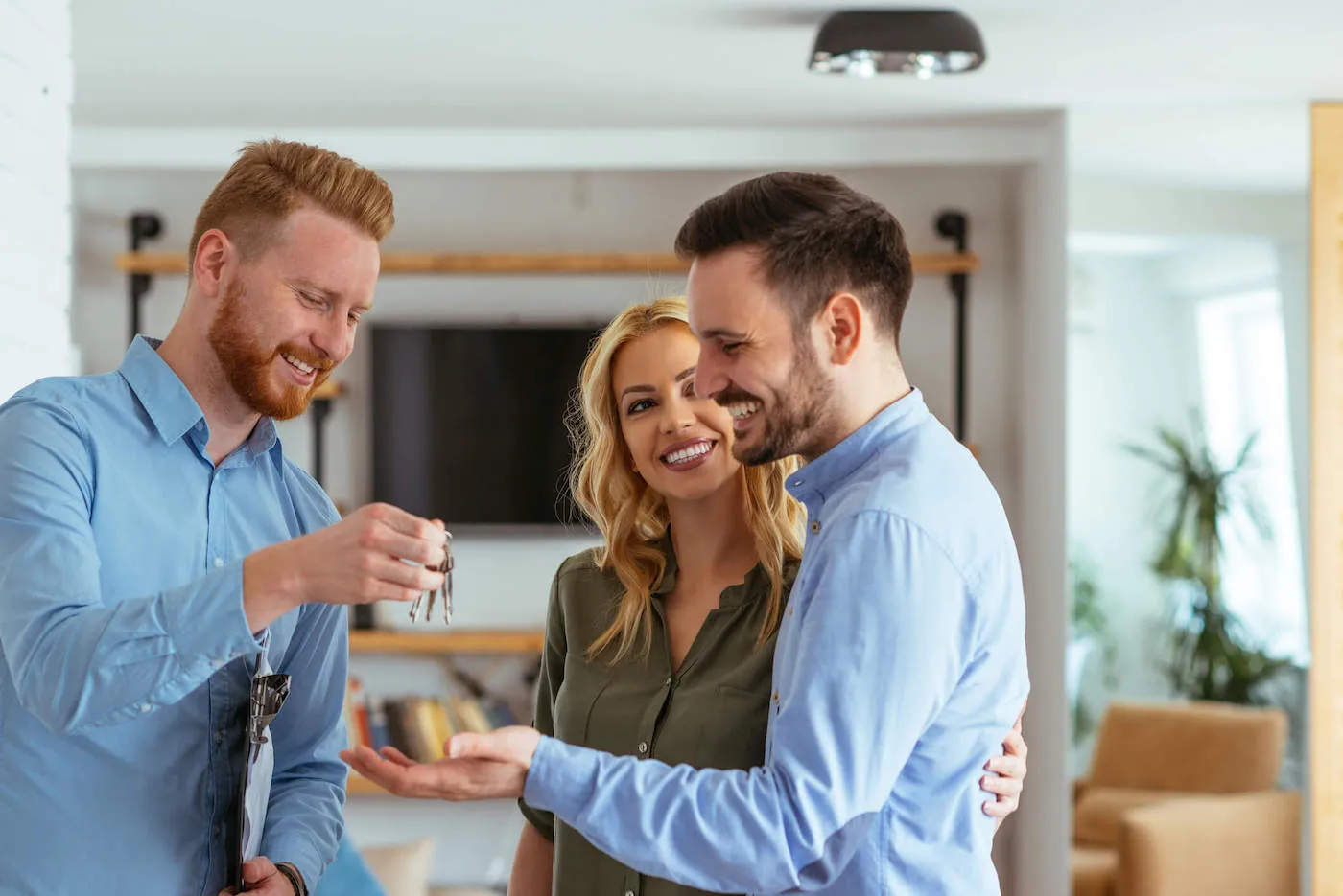 A man wearing a blue shirt smiles as he gives a set of house keys to a couple.