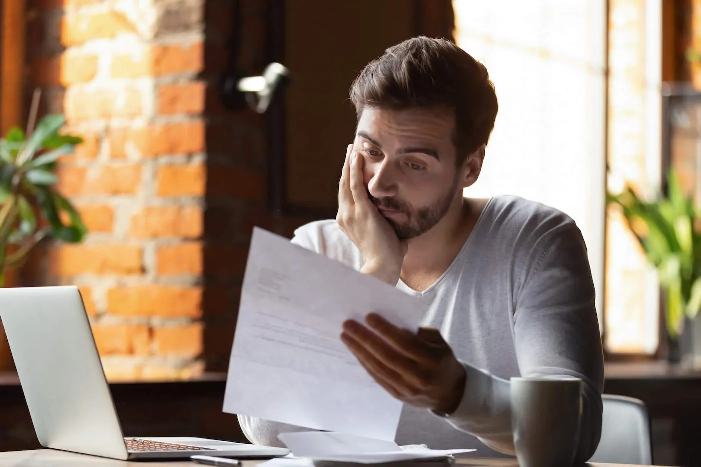 A man wearing a gray shirt is leaning on his hand has he looks at a document with his laptop open on the table.