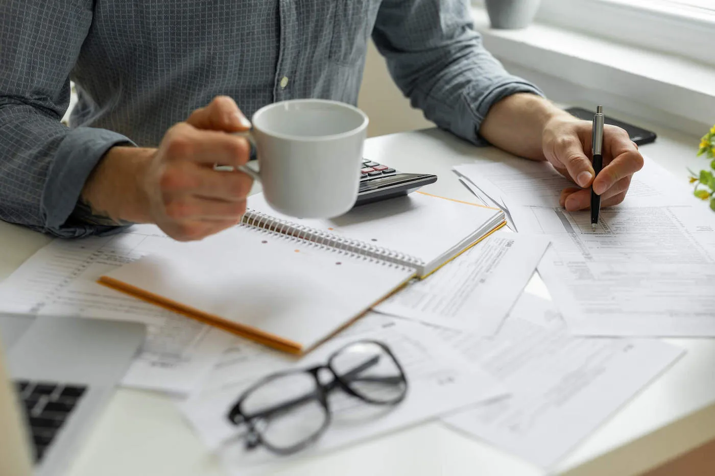 A person holding a coffee cup writes on tax form documents with a calculator, laptop, notebook, and reading glasses on the table.