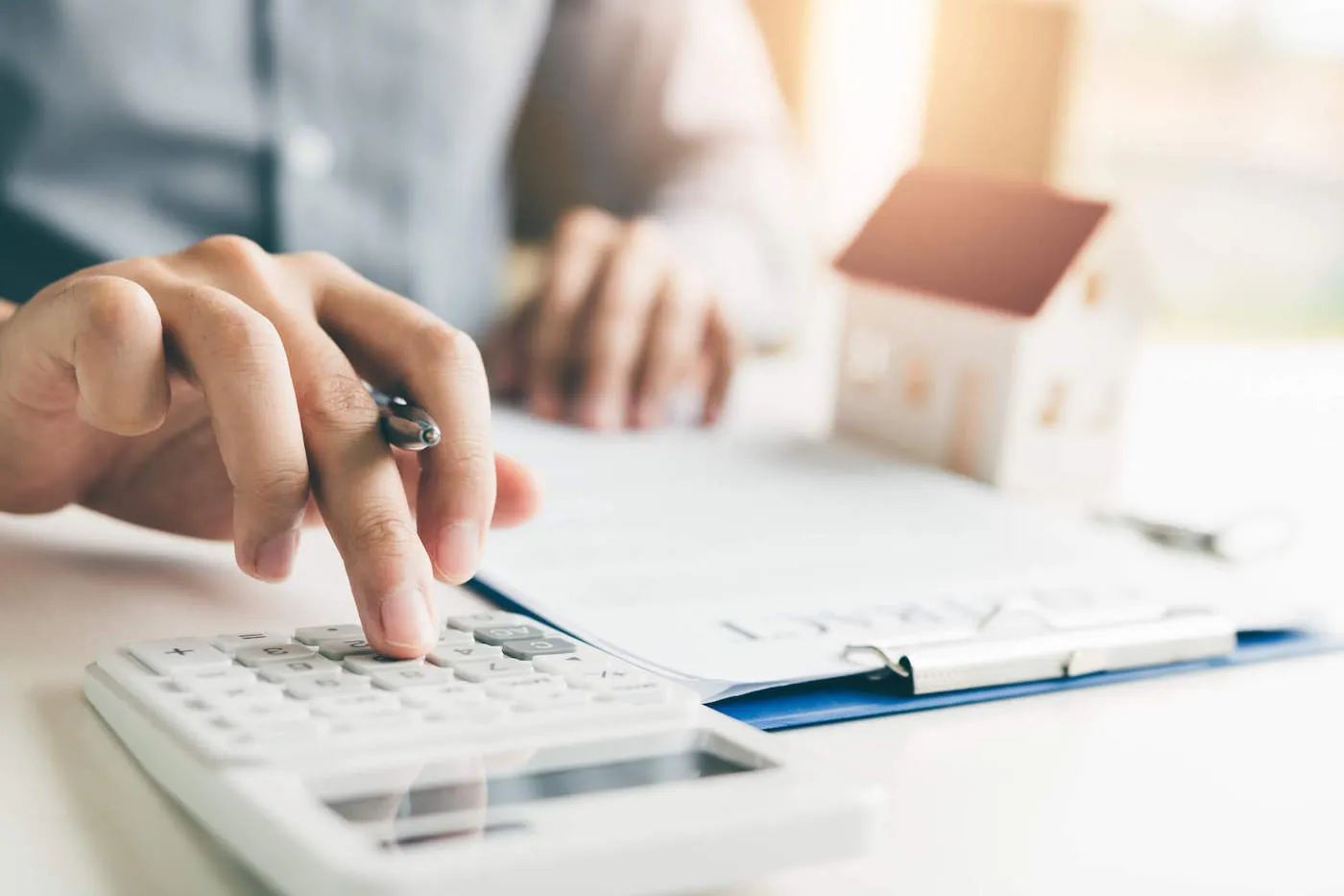 A person uses a calculator while looking at a document with a miniature house model next to it.