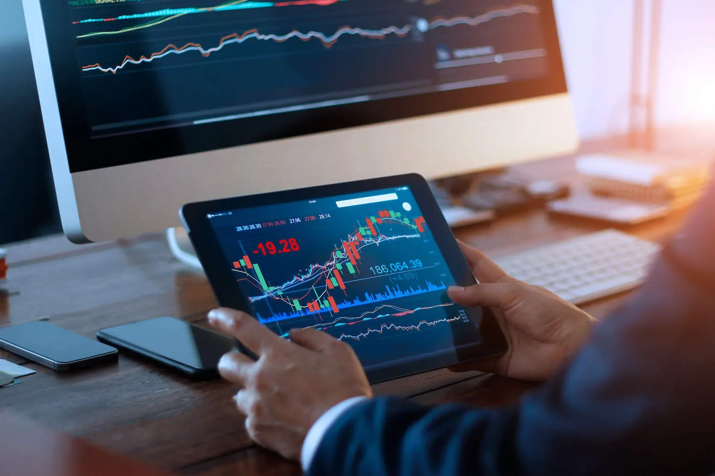 A person wearing a navy blue suit looks at a stock graph on their tablet screen with their desktop computer in the background.