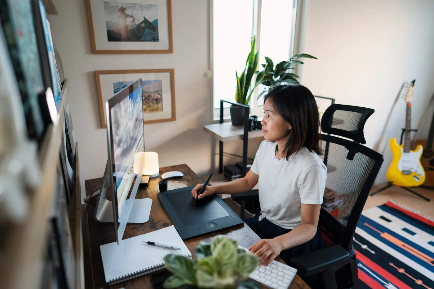 A woman looks at her computer while drawing on a digital trackpad at home.