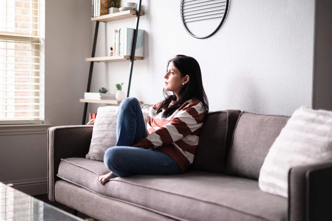 A woman frowns as she looks out the window while sitting down on the couch.