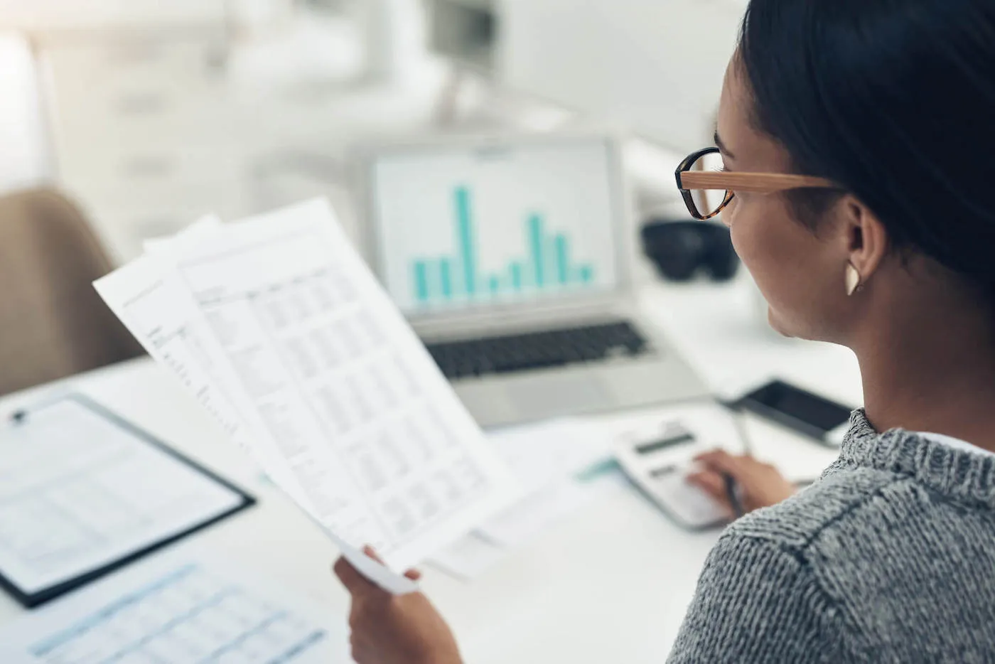 A woman wearing glasses looks at documents while using a calculator and laptop at her work desk.