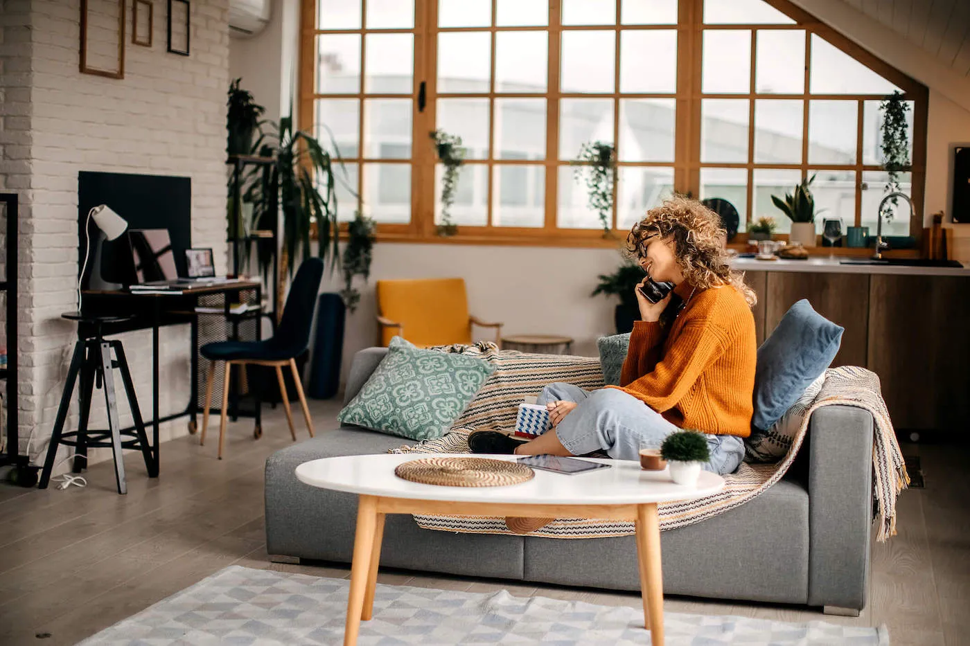 A woman wearing an orange sweater sits on her couch while she talks on the phone inside her condo.