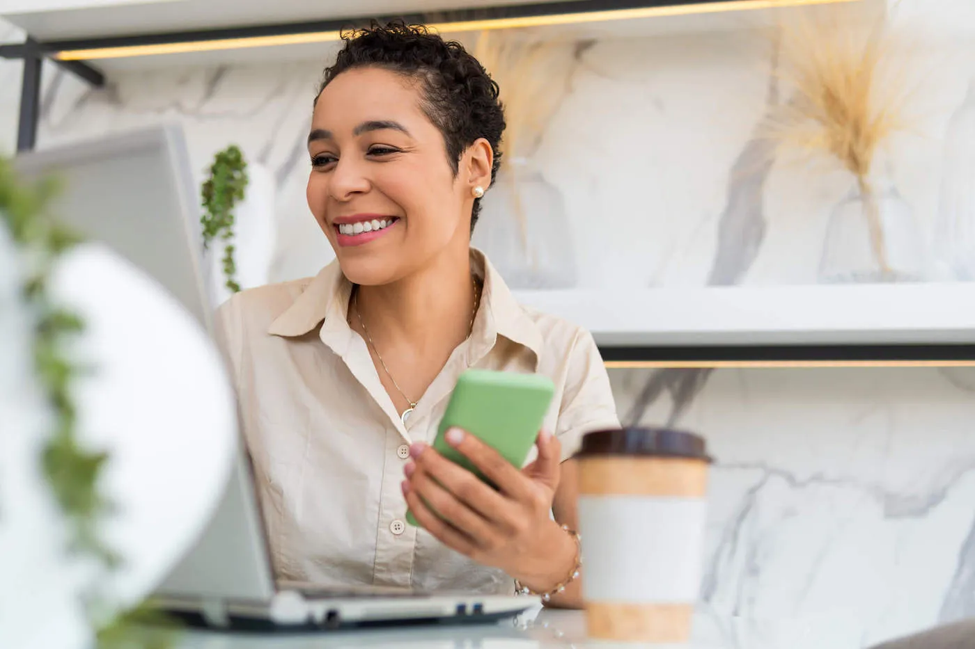 A woman smiles at her laptop screen while holding her green phone.