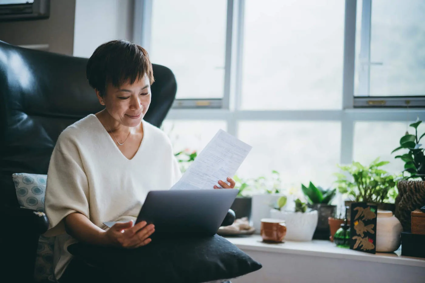 A woman is sitting down while smiling at her laptop as she holds a document.