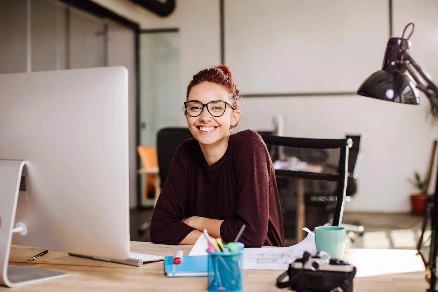 A woman with red hair is smiling at the camera while in front of her work desk.