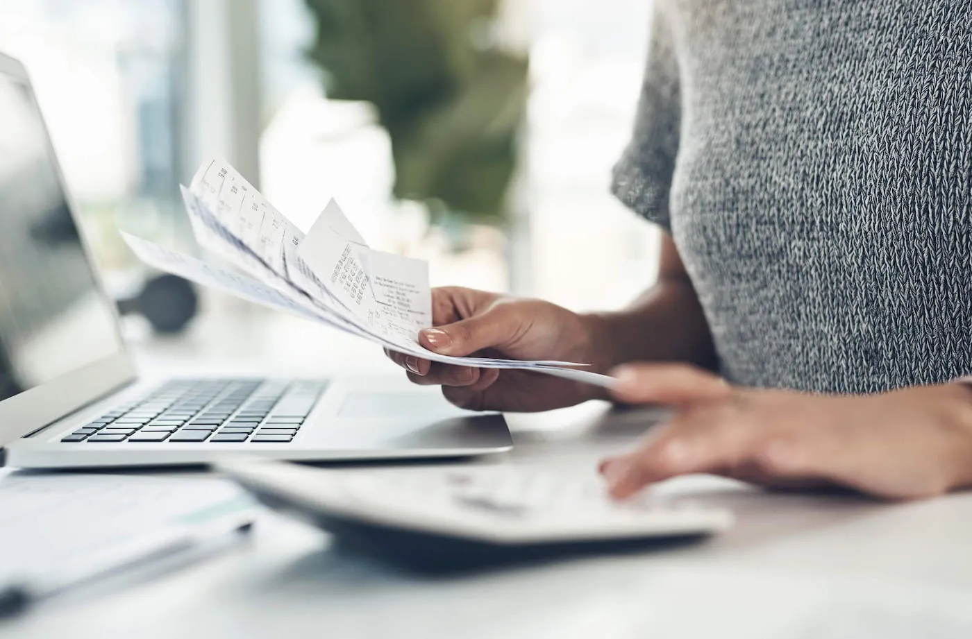 A woman wearing a gray sweater uses a calculator while looking at receipts.