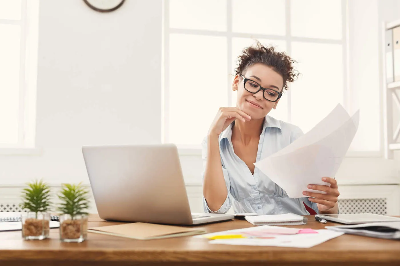 A woman wearing a blue shirt looks at documents with her laptop on her work desk.