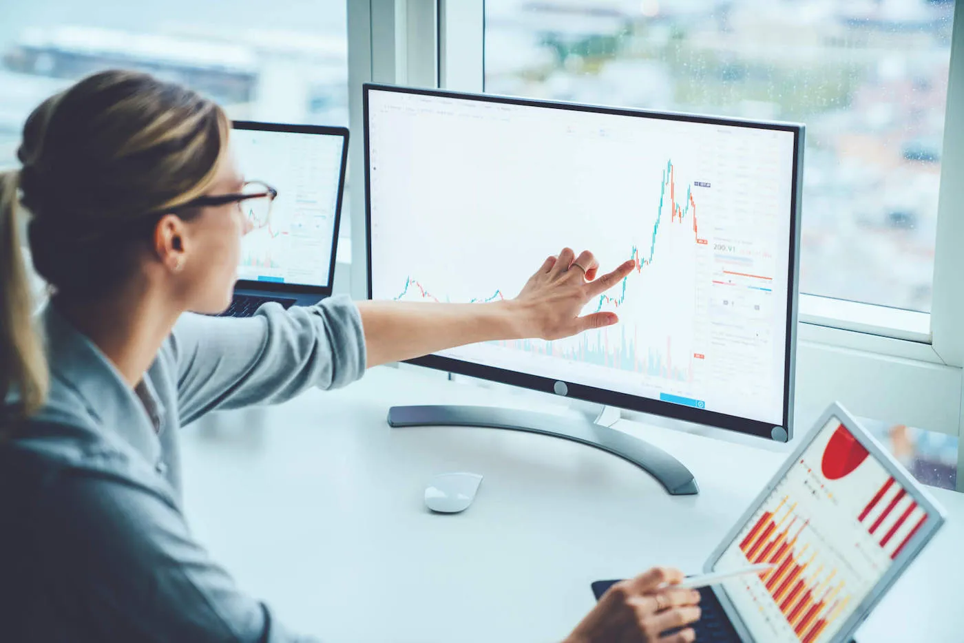 A women with blonde hair is pointing at a stock graph on her computer screen as she uses an iPad.