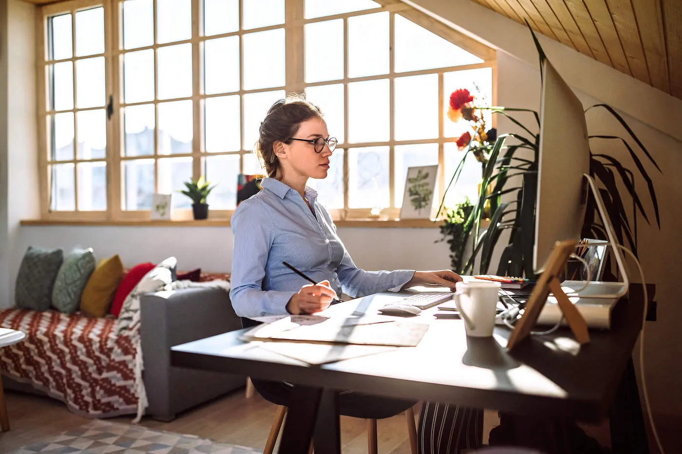 A woman wearing glasses holds a pencil while looking at her computer from home.