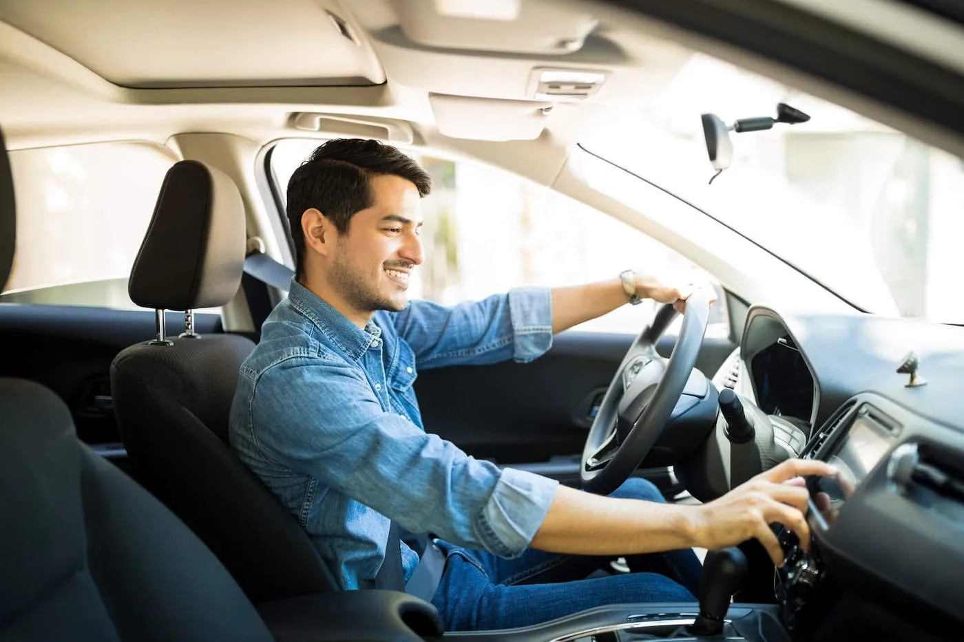 A young man wearing a denim shirt smiles while touching his car radio.