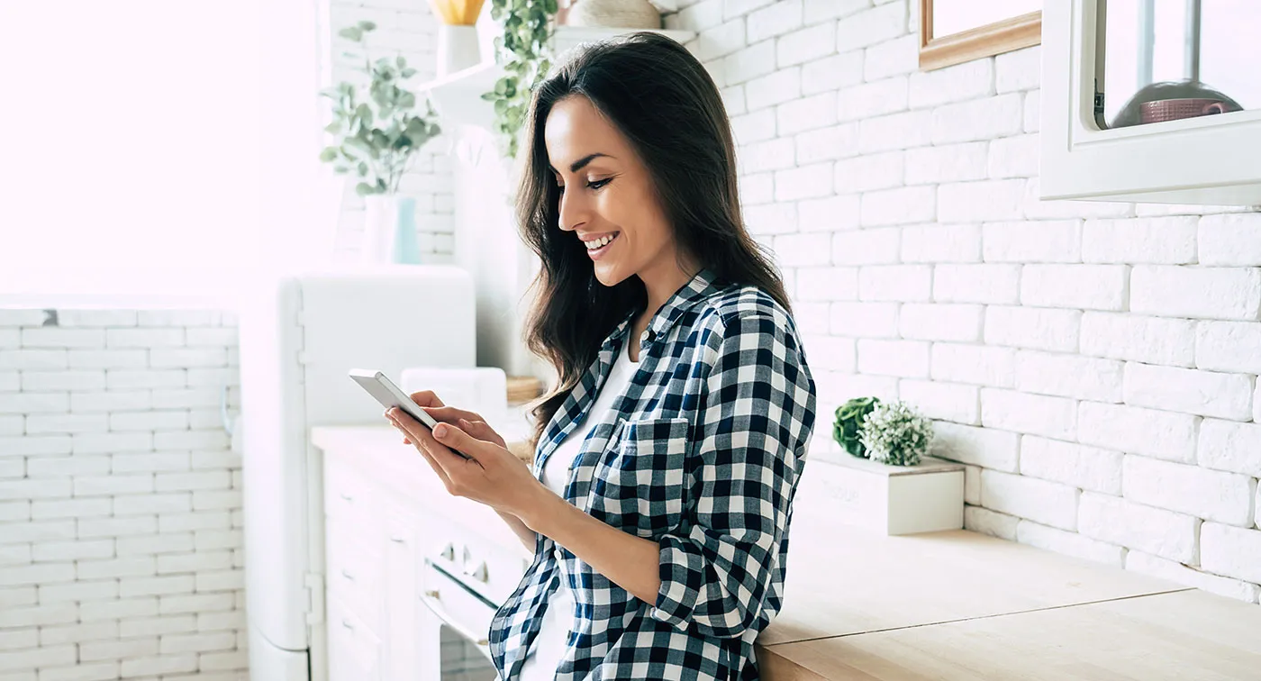 A woman wearing a flannel shirt smiles at her phone while she's in the kitchen.