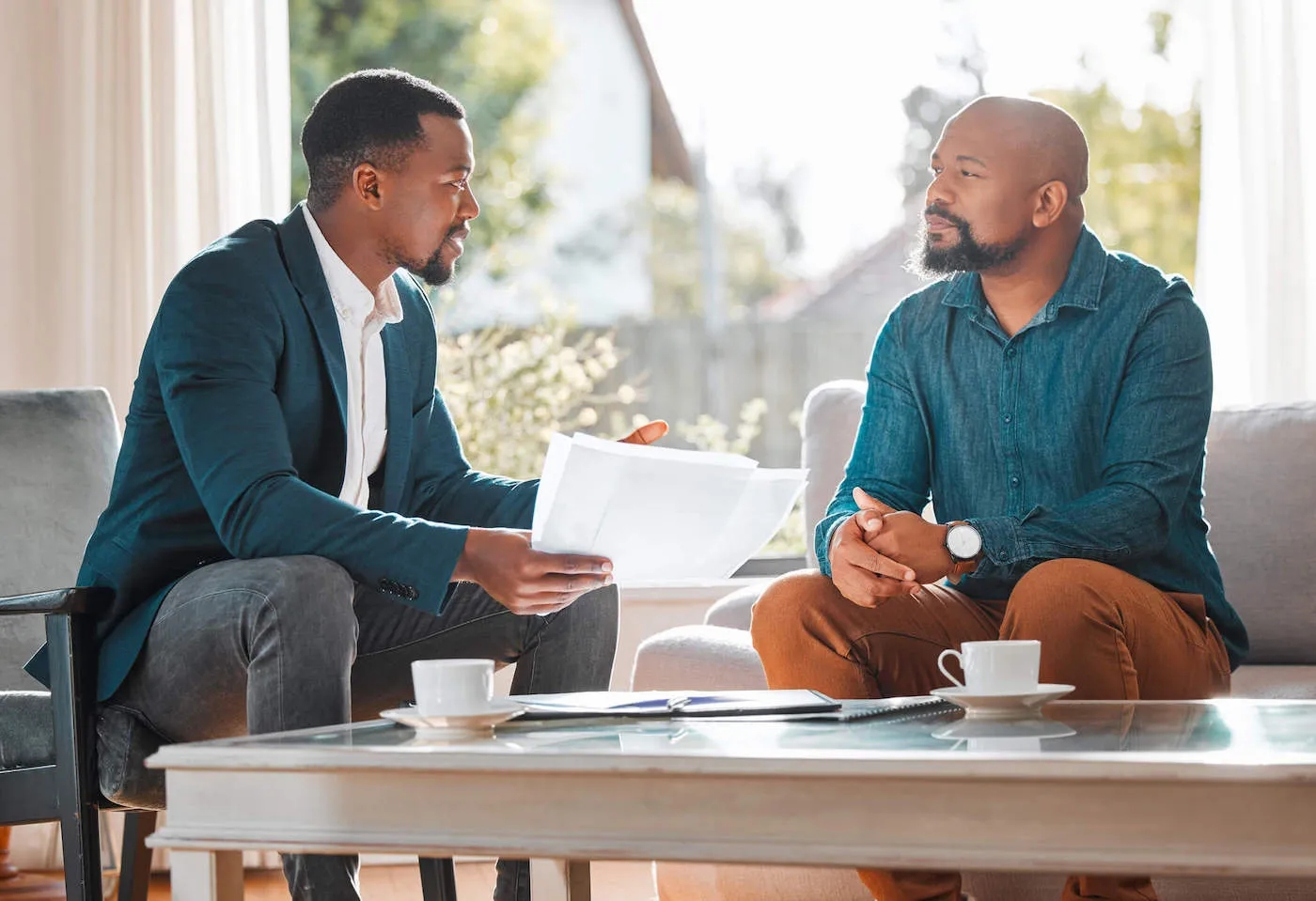 An agent wearing a blue suit is talking to his client at the living room of a home while showing him documents.