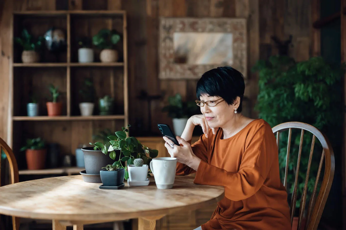 An elderly woman wearing an orange shirt sits down while looking at her phone with plants in the background.