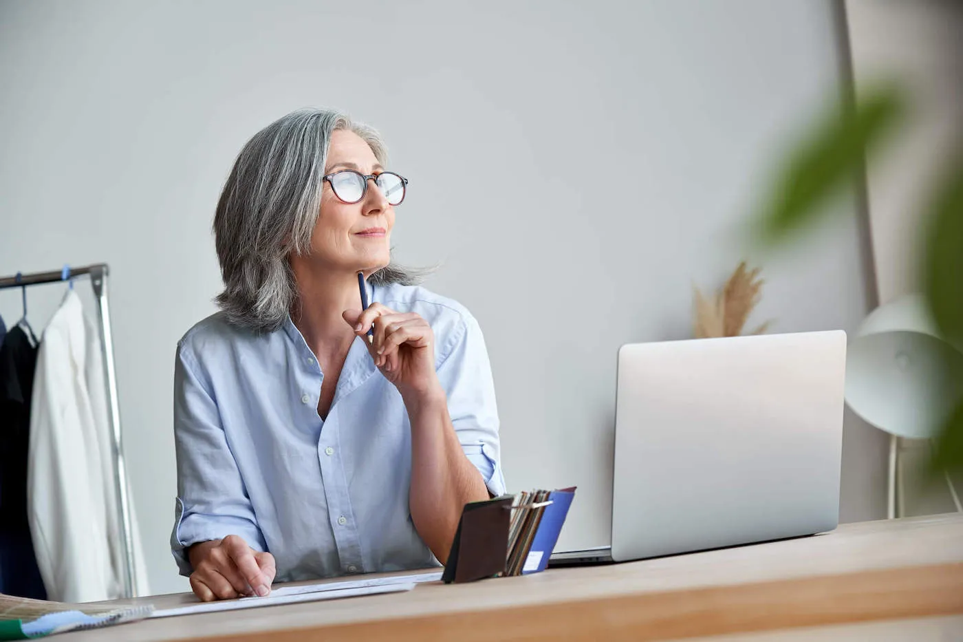 An older woman wearing glasses looks outside the window and smiles while her laptop is next to her.