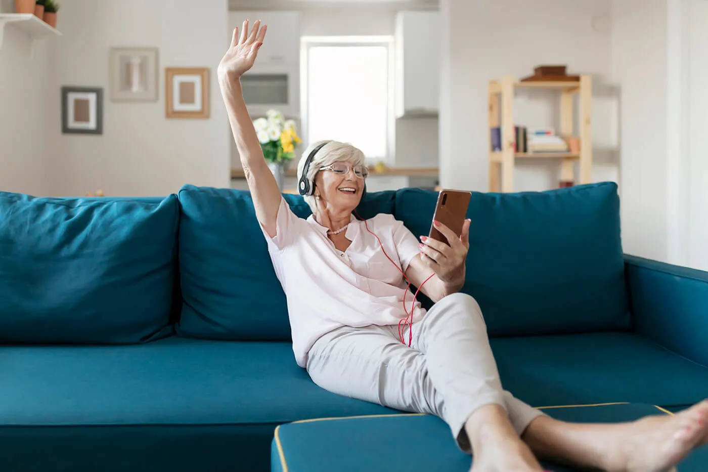 Cheerful senior woman with headphones listening to the music and using her smartphone while relaxing on the couch