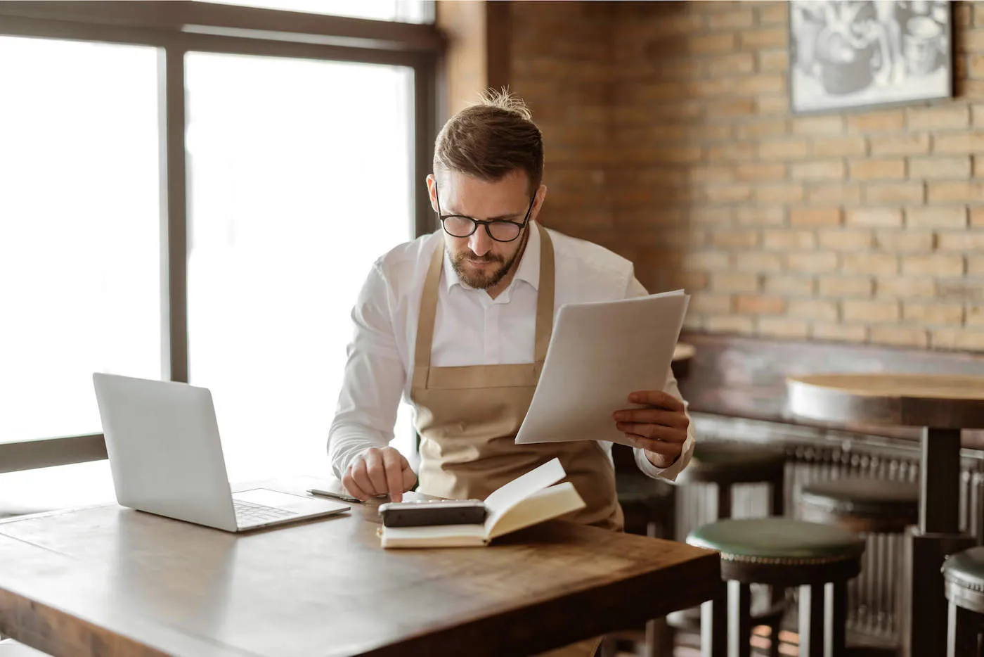 A business owner wearing glasses and a brown apron holds documents while using a calculator at a restaurant.