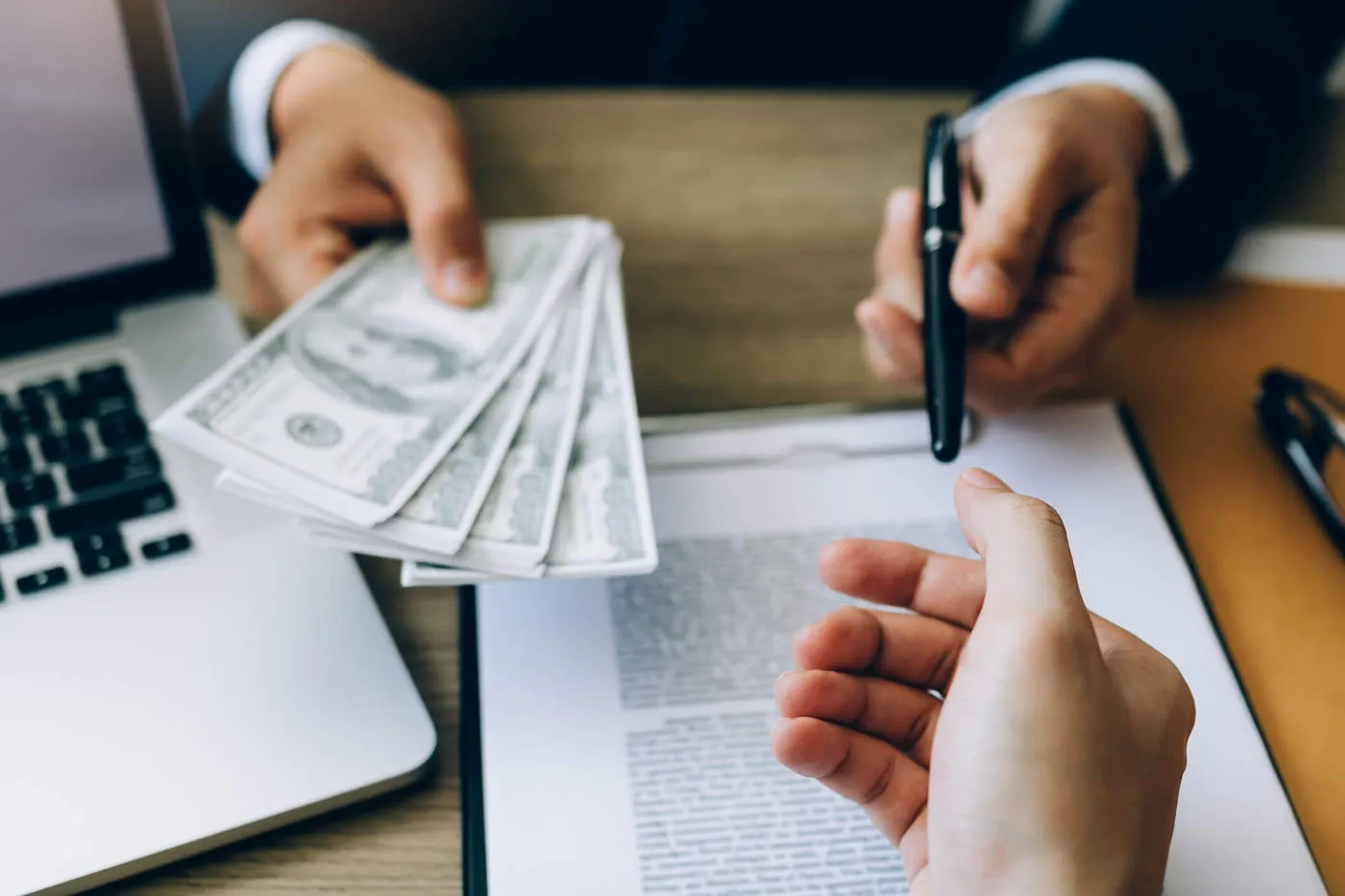 A closeup shot of a person's hands giving another person cash and a pen to sign a document on the table.