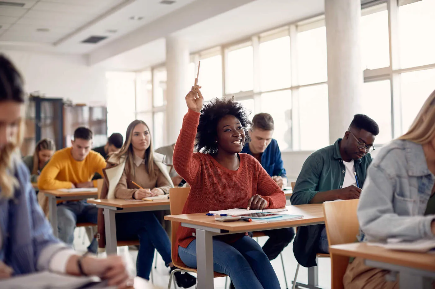 A group of college students are in a classroom and a student wearing a red sweater is raising her hand.