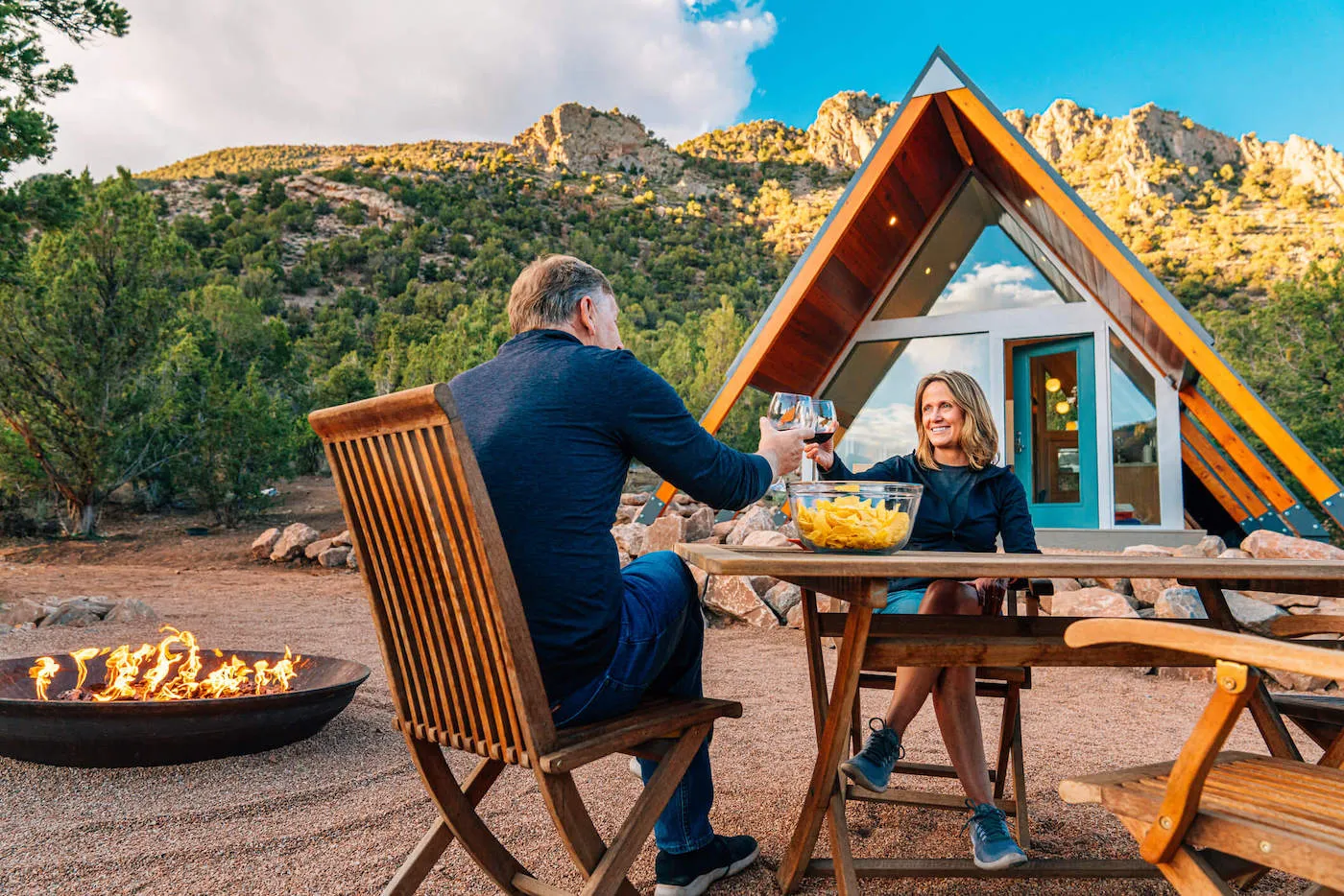 A couple smile and cheers each other with their wine glasses in front of their home with a mountain in the background.