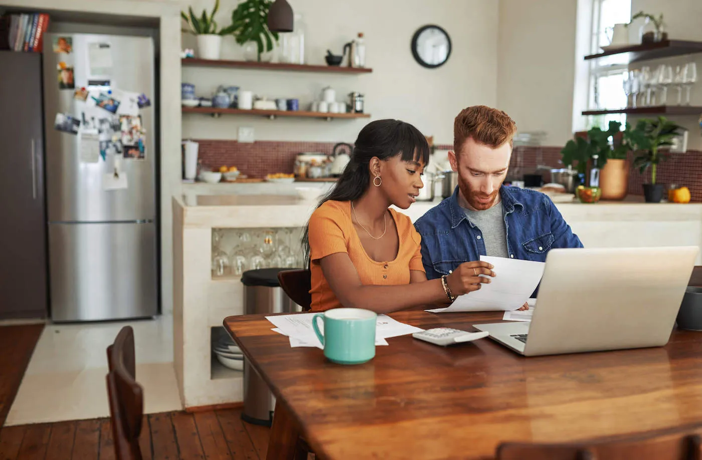 A couple at the kitchen table sit next to each other while looking at documents.