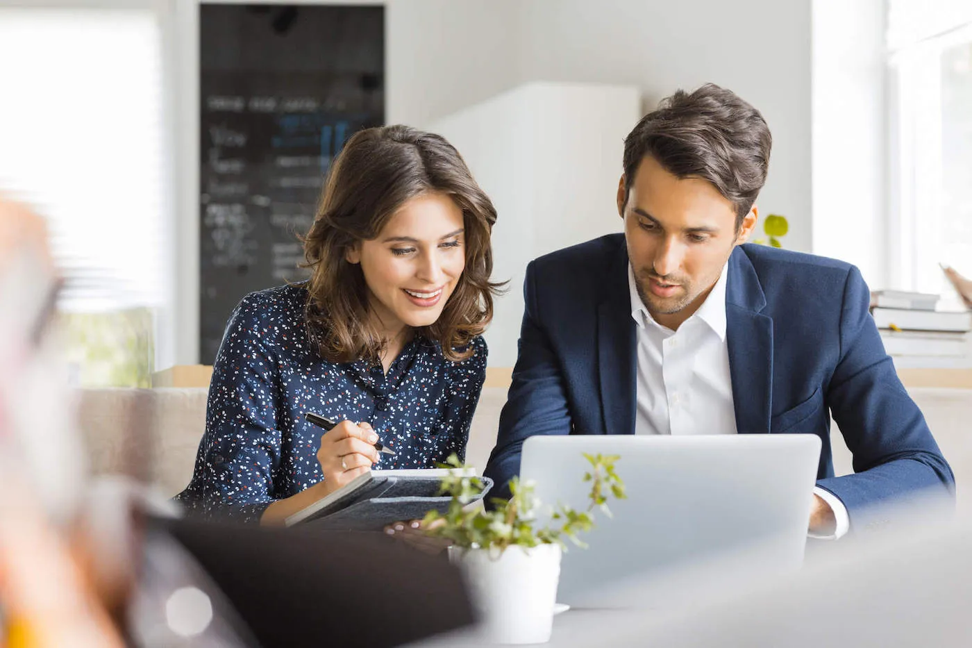 A woman takes notes on her notebook as she sits next a man wearing a suit and using a computer.