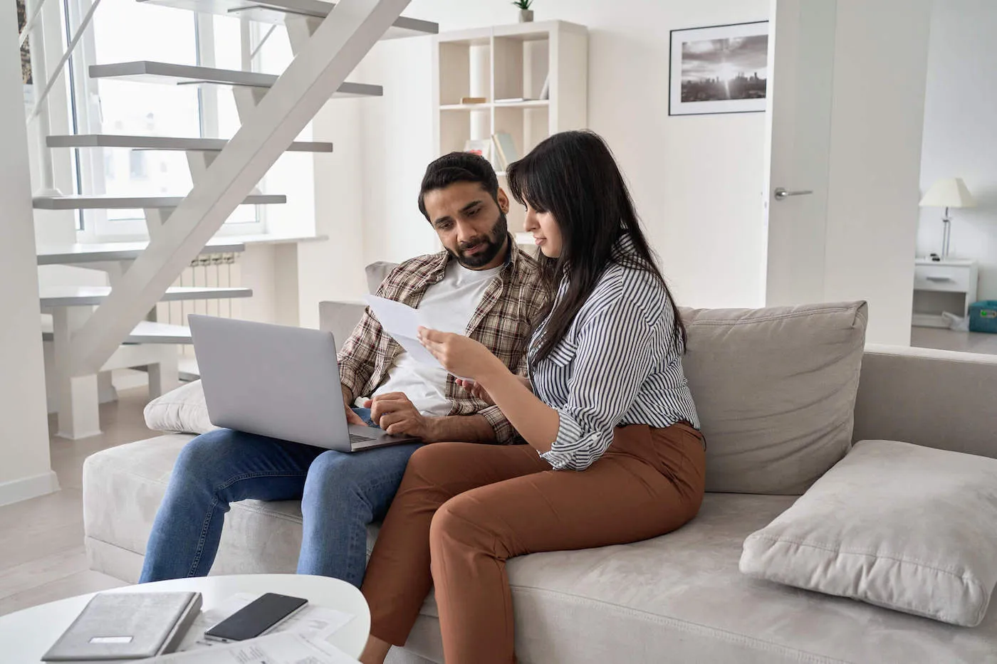 A couple sitting on the couch together looks at a document while the man uses his laptop.