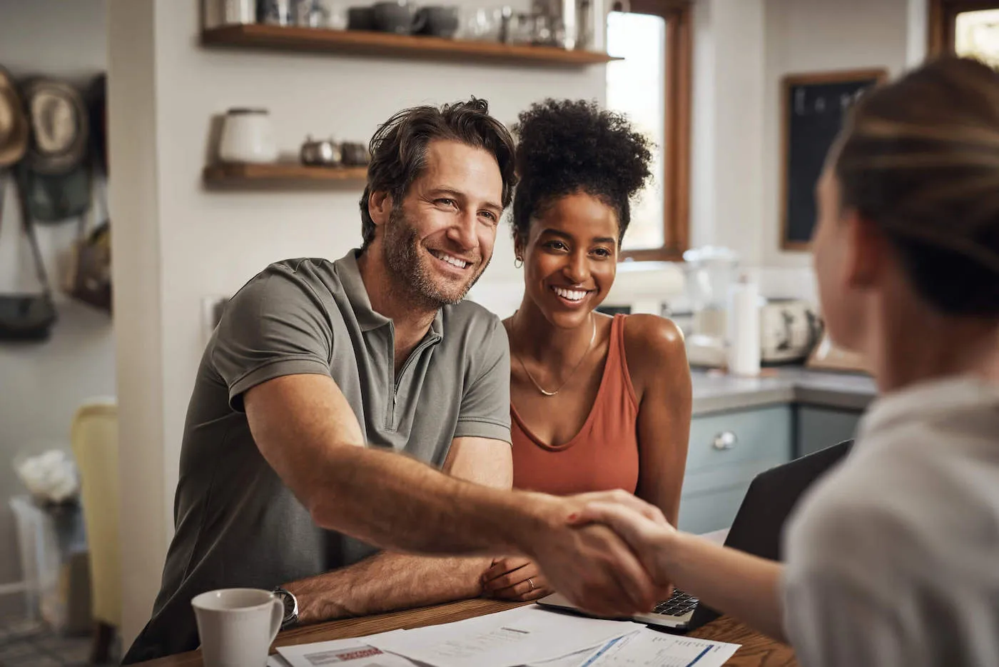 A couple smile at an advisor from across the table and the man shakes her hand with documents on the kitchen table.