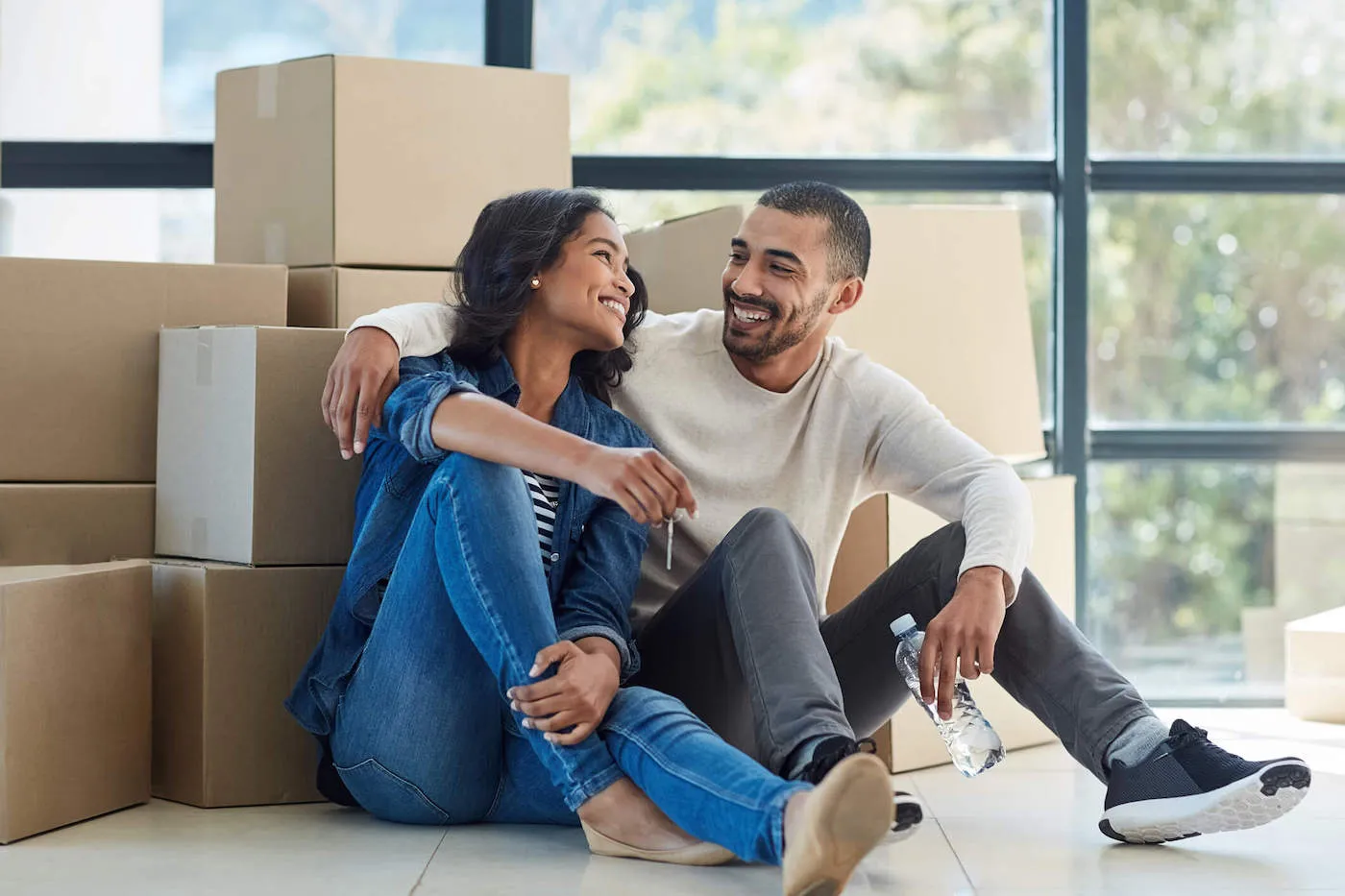 A couple smiles at each other as they sit on the floor of their new home while they have moving boxes behind them.