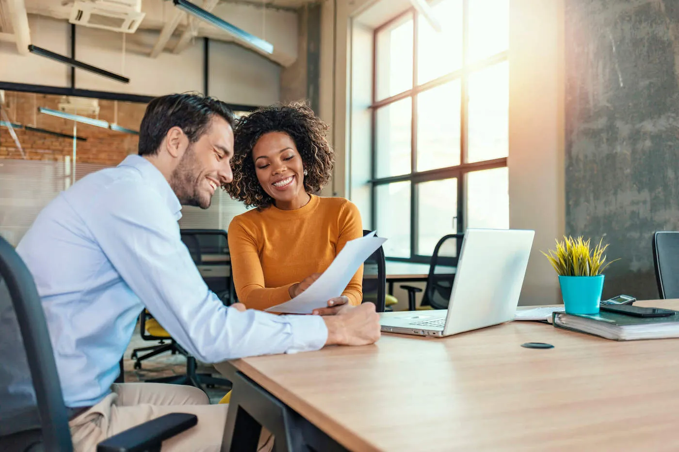 Two coworkers are smiling while looking at a document with a laptop on the table at the office.