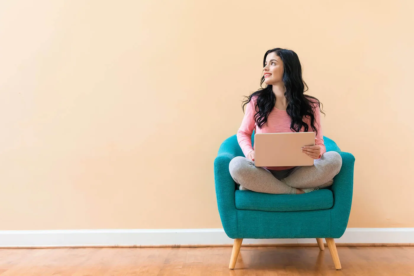 A smiling woman with crossed legs sitting on a turquoise armchair and holding a laptop