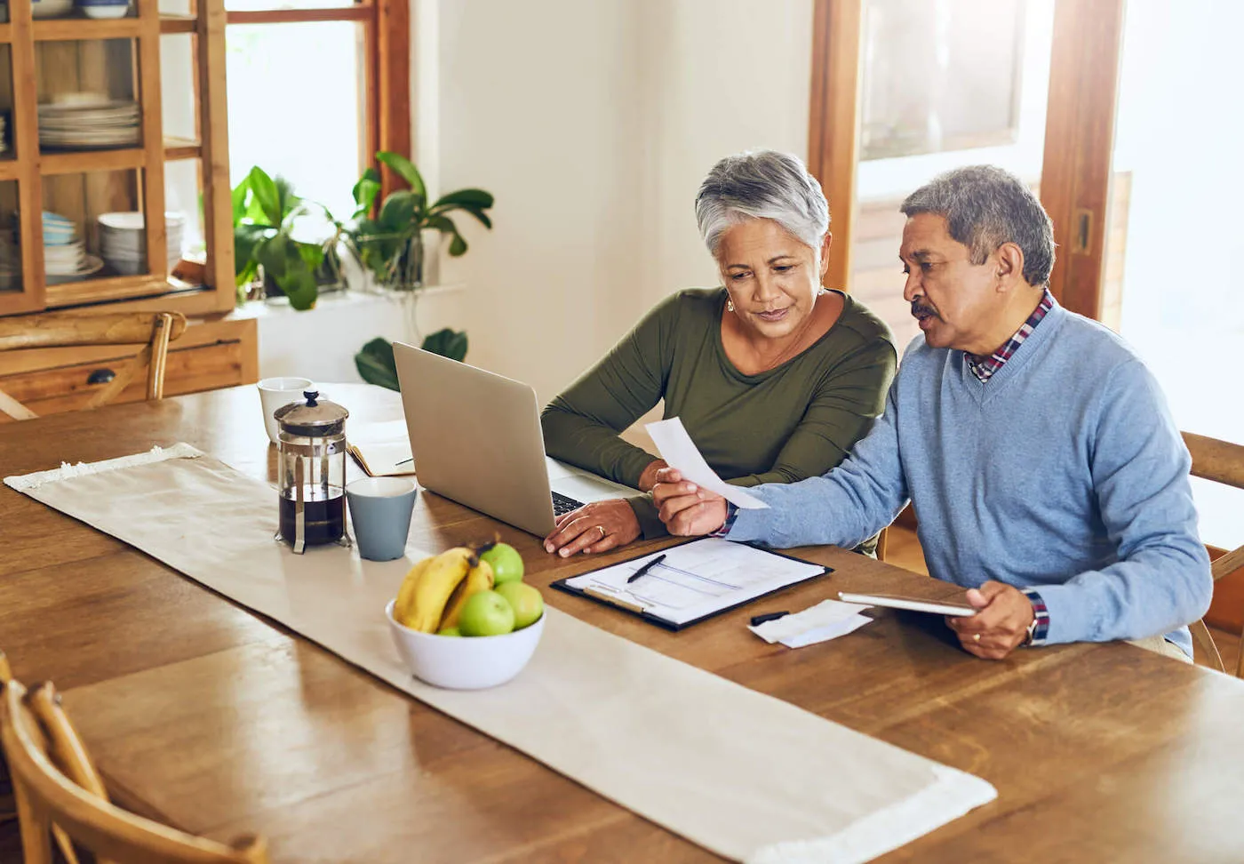 An elderly couple look at a document while their laptop is on the kitchen table.