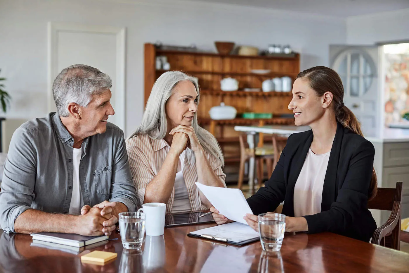 An elderly couple talk to a woman wearing a suit at the living room table.